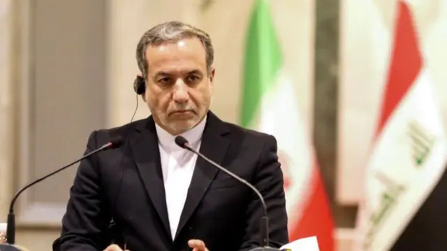 Iranian Foreign Minister Abbas Araghchi standing at a lectern with two microphones in front of him and an earpiece in