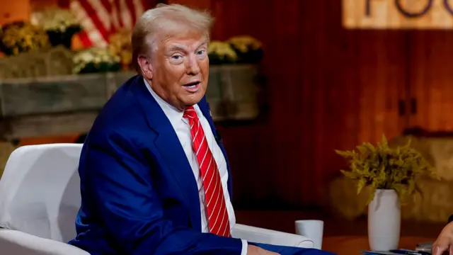 Donald Trump sitting on a television set wearing a navy suit and red tie