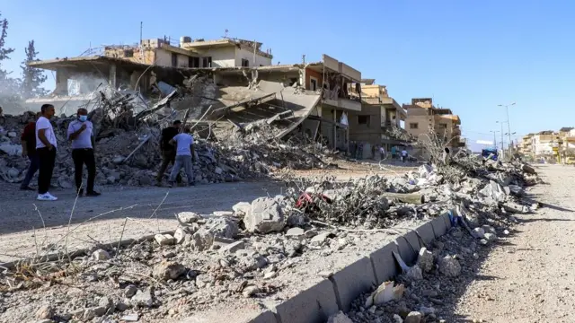 A house on the side of the road is reduced to rubble, as men - including one wearing a face mask - stand and look at the damage