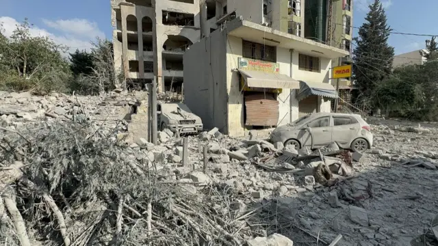 A view of cars covered with thick grey dust and ash, next to debris and a collapsed building.