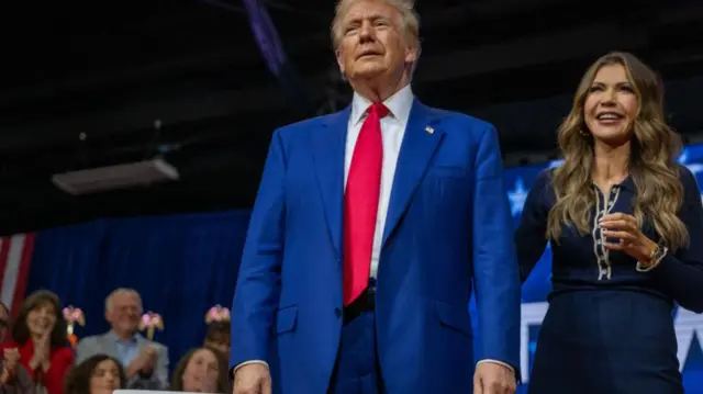 Trump stands onstage, flanked by a brunette woman, South Dakota Governor Kristi Noem