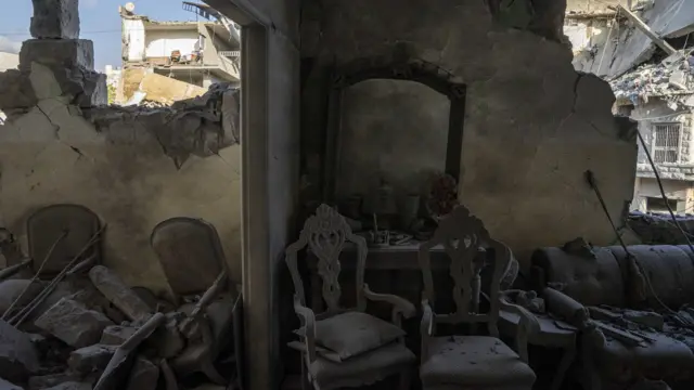 The inside of a building that was bombed in an air strike, it shows ornate chairs and a sofa covered in thick dust and debris