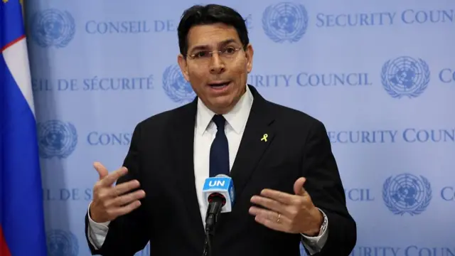 Danny Danon wears a black suit and blue tie as he stands at a podium and speaks into a microphone with UN branding. The wall behind him reads: UN Security Council