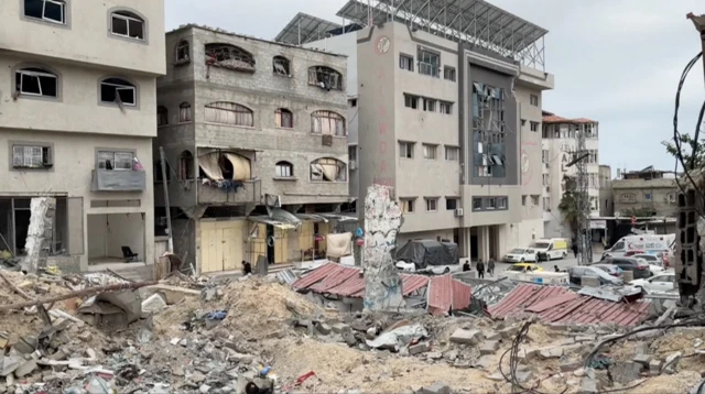 Damaged facade of al awda hospital in Jabalia. Debris are piled at front of damaged grey building with broken windows. Parking lot in the background