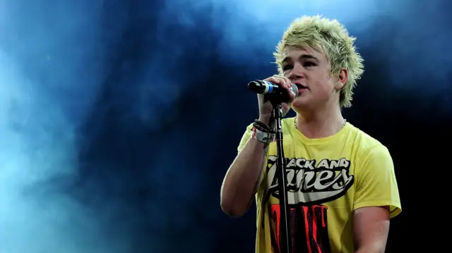 Eoghan Quigg performs at the Isle Of Man Bay Festival in June 2010. He is wearing a yellow shirt, had bleached-blonde hair and is standing on a stage covered with smoke