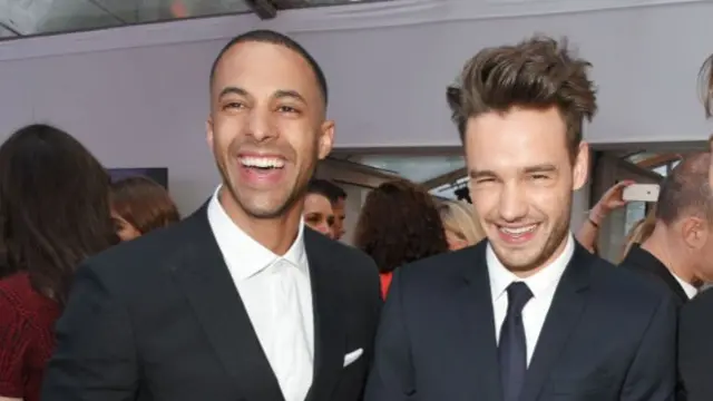 Marvin Humes in dark suit and white shirt smiles at camera. Liam Payne in dark suit, black tie and white shirt stands next to him also smiling