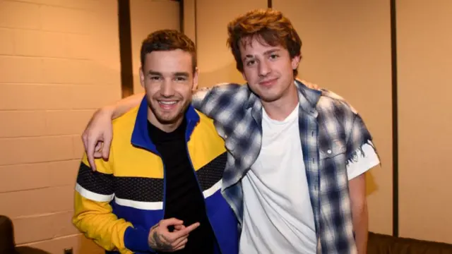 Liam Payne in black shirt and blue, black and yellow striped jacket with arm around Charlie Puth's shoulder. Puth is wearing a sleeveless white t-shirt and plaid shirt