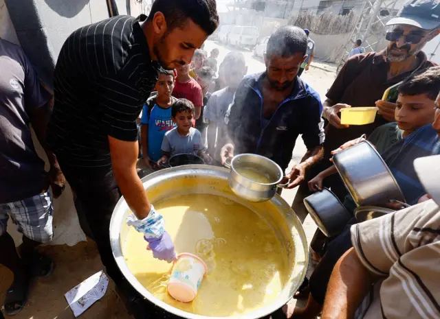 A man scoops soup from a large saucepan as people crowd around with containers