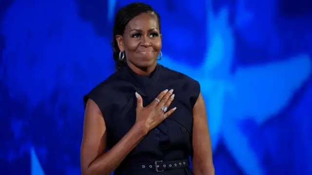 Obama looks out, with her hand over her heart, as she stands in front of a blue background