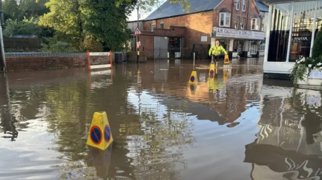 A flooded street with yellow cones standing in the water and a man with a yellow jacket walking through the flood