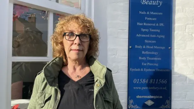 A woman with blonde curly hair and a green jacket standing in front of a white building with a white-framed window. window and a blue sign