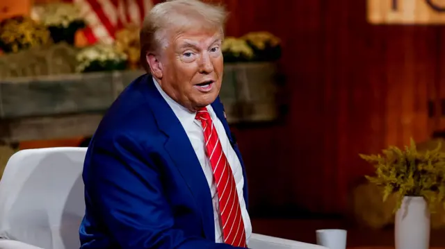 Trump, wearing a blue suit and red striped tie, leans forward while sitting on a white chair