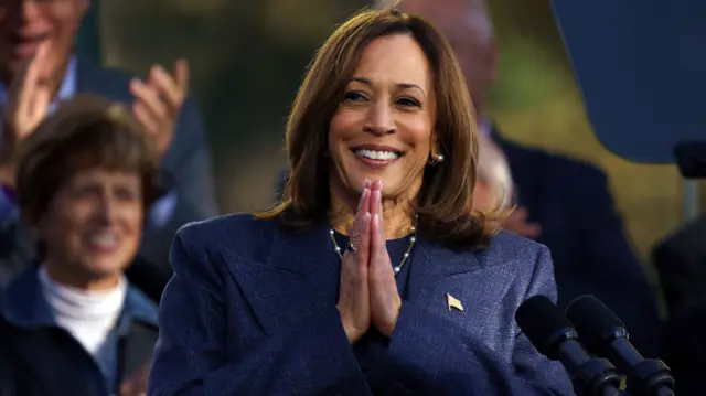 Kamala Harris, wearing a blue suit, places her hands together across her chest during a campaign event