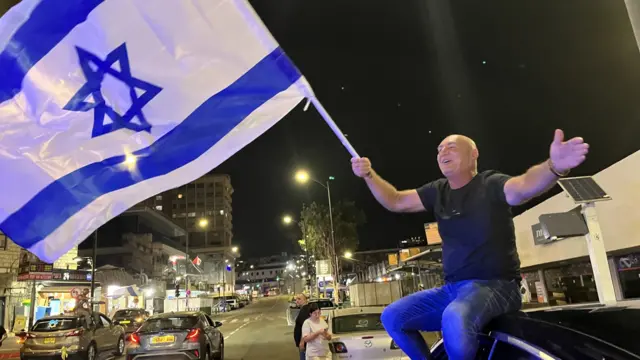 A man sits on a car smiling and waving an Israeli flag