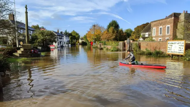 A paddle-boarder on a flooded street. Brown floodwater dominates the scene with a war memorial and phone box visible