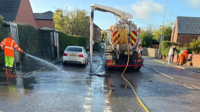 A man hosing down a street with a lorry with a tank on the back supplying water