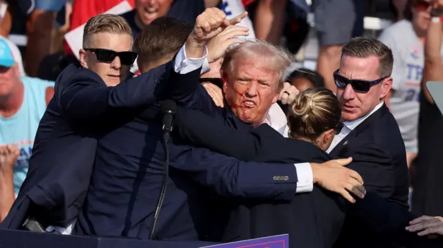 Donald Trump, with blood on his face, is escorted off a stage by US Secret Service agents