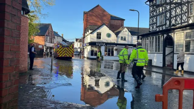 A fire engine and several men in yellow high-vis jackets in a street with a black-and-white timbered building on one side and red brick buildings on the other and there is water lying on the road