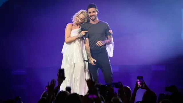 Rita Ora, wearing a long white dress, leans on Liam Payne's shoulder on stage as mobile phones are held up in front of them