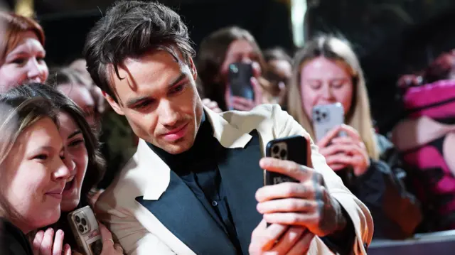 Liam Pain, a dark haired man wearing a white suit with black frontage, takes selfies with female fans