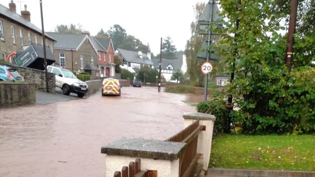 A street with a garden on the right hand side and brown water covering the street and church in the distance