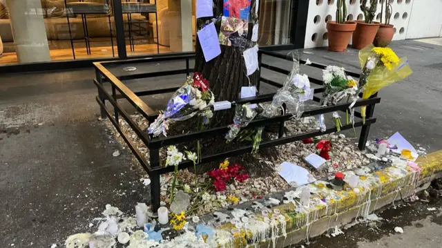 Flowers and melted candles lay in tribute to Liam Payne by a tree outside a hotel