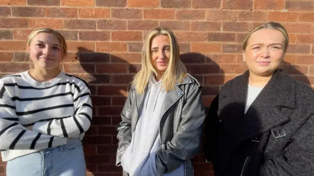 Lilah Rose in striped jumper and jeans, Faith in black leather jacket and grey hoodie and Caity in black jacket and grey t-shirt stand in a row against a brick wall in Wovlerhampton