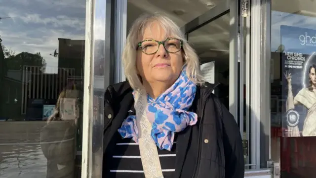 A woman with blonde hair, black coat and blue and pink scarf standing in a doorway