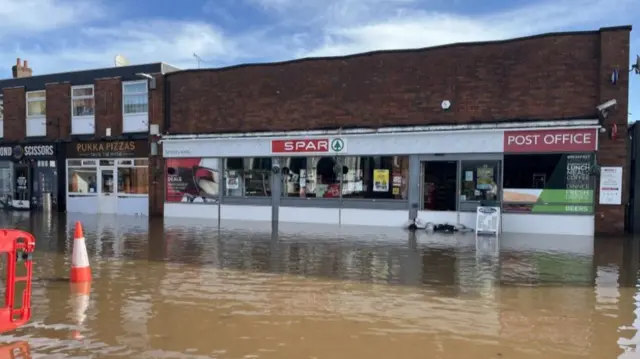 A Spar shop with brown water covering the road in front of it and a red cone sitting in the water