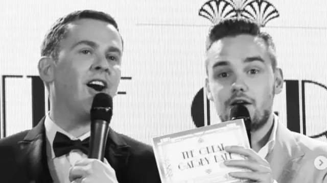 Black and white picture of Scott Mills wearing a tuxed and holding a mic standing next to Liam Payne. Payne is wearing a suite and white shirt and holding a mic and card reading The Great Gatsby Ball in his left hand