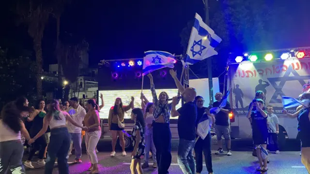 People dance and raise their arms, three are waving Israeli flags