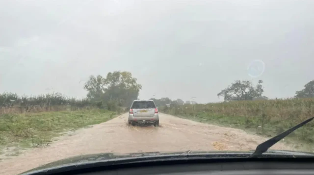 A view through a car windscreen of water on a road with grass on either side and a silver car in front driving through the brown water