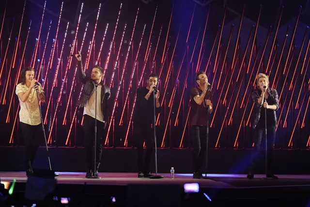 The five members of One Direction are seen on stage in front of an orange and red backdrop.