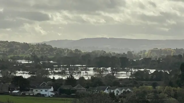 Aerial shot showing houses in the foreground with flooded fields behind against a backdrop of hills and trees