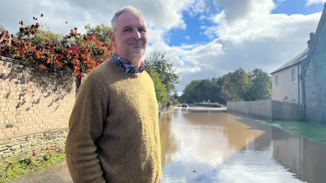 A man in a floral shirt and brown jumper stands at the edge of floodwater outside a house. In the distance, a stranded car is visible in the water.