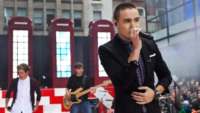 Liam Payne, sings into a microphone in front of bandmates