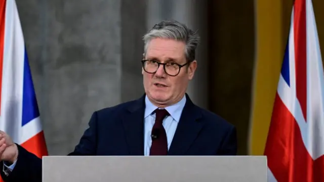 Keir Starmer in black suit with matching black tie and powder blue shirt. He's wearing glasses as he delivers speech in Berlin. Two UK flags stand at his sides