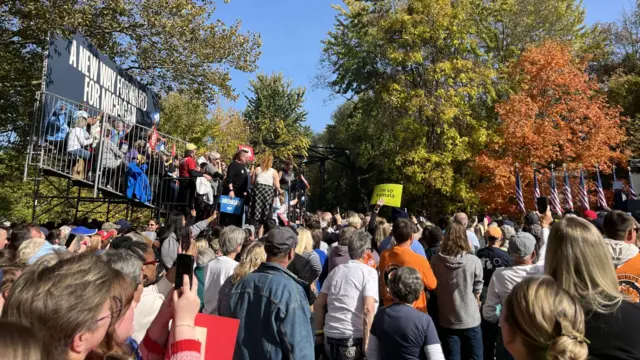 A crowd of people gather outdoors for a Kamala Harris rally on a sunny day surrounded by trees