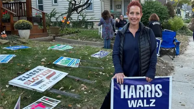 Marcie Paul holding a Harris Walz sign in front of a lawn with lots of other signs