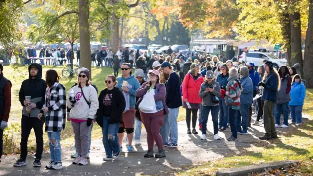 People line up on a path surrounded by trees and greenery. The line is very long and curves around. It is about 2 to 4 people deep