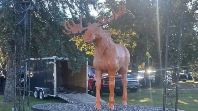 A large orange statue of a moose surrounded by evergreen trees, with the sun peeking through the branches