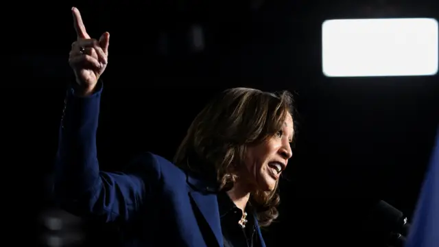 Kamala Harris, wearing a blue suit, points to her right while addressing a crowd