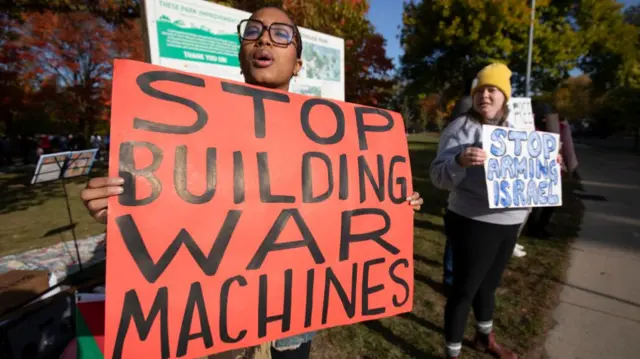 A black woman holding a sign that reads stop building war machines. A white woman stands next to her with a sign that reads stop arming Israel. Autumnal trees can be seen in the background