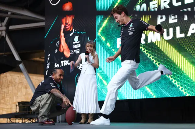 George Russell kicks an American football, which is being held by Lewis Hamilton on stage