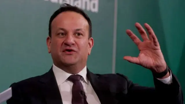 Leo has short black hair. He has his hand up in the air and is mid conversation. He is speaking against a green background. He is wearing a white shirt and maroon tie.