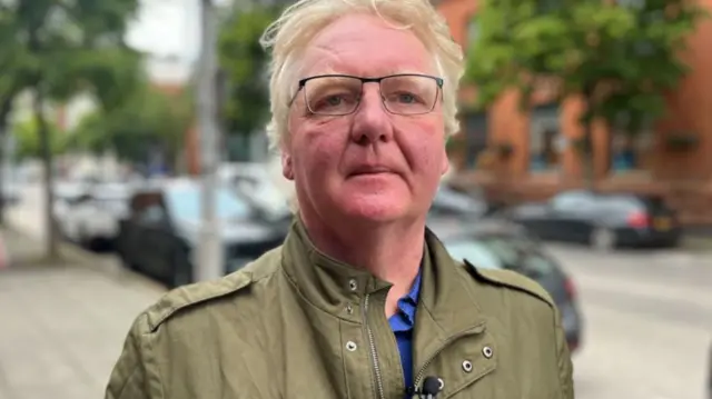 James is standing in front of a street with parked cars and lamposts. He has greyish strawberry blond hair and eyebrows and wears square thin black glasses. He is looking directly at the camera and is wearing a green jacket and blue shirt underneath.