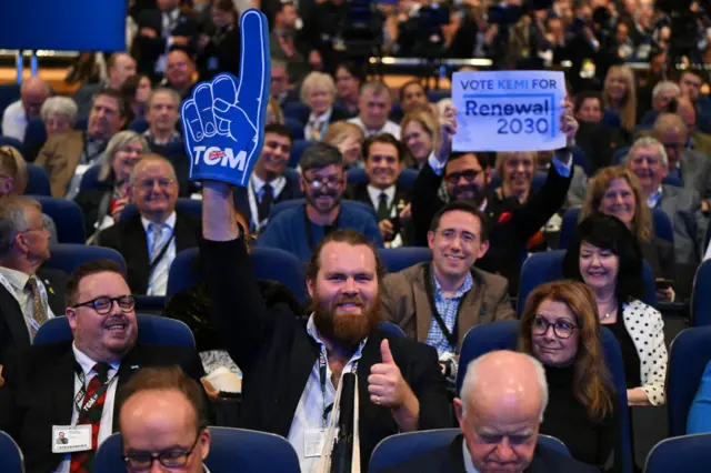 Conservative voters sitting during conference. One man in dark suit with thumbs up and arm raised with foam finger