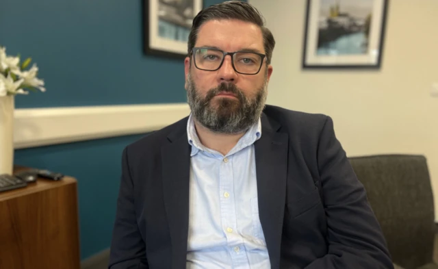 A man with short, dark hair wearing square framed glasses sits in a blue and white wall room. He is looking at the camera and wearing a dark suit jacket and blue collared shirt.