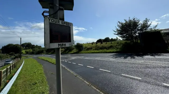 A main road with single white line markings and two lanes. There is a sqaure shaped speed detector with the words 'YOUR SPEED MPH' written in capitals. The sky behind is blue and sunny with a few clouds. The road is lined by grass and some trees.