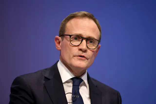 Tom Tugendhat mid-speaking on stage wearing black suit with white, wide-collared blouse and dark blue tie